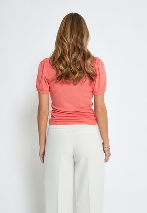 Koralo spalvos trumpomis rankovėmis marškiniai, prigludusi siluetas. Derinami su šviesiai kreminių plačių kojų kelnėmis, turinčiomis lygią tekstūrą ir užpakalines kišenes.