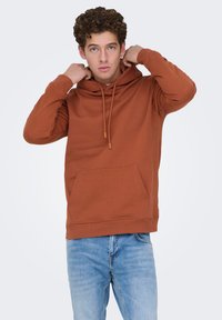 Bruine trui met een voorzak en een capuchon met trekkoord. Zachte stof, casual pasvorm, geribbelde manchetten en zoom. Draagt bij lichtblauwe spijkerbroek.