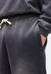 Κοντινό πλάνο προσώπου που φοράει σκούρο γκρι φόρμα με λευκό κορδόνι και το χέρι κρυμμένο στην δεξιά τσέπη.