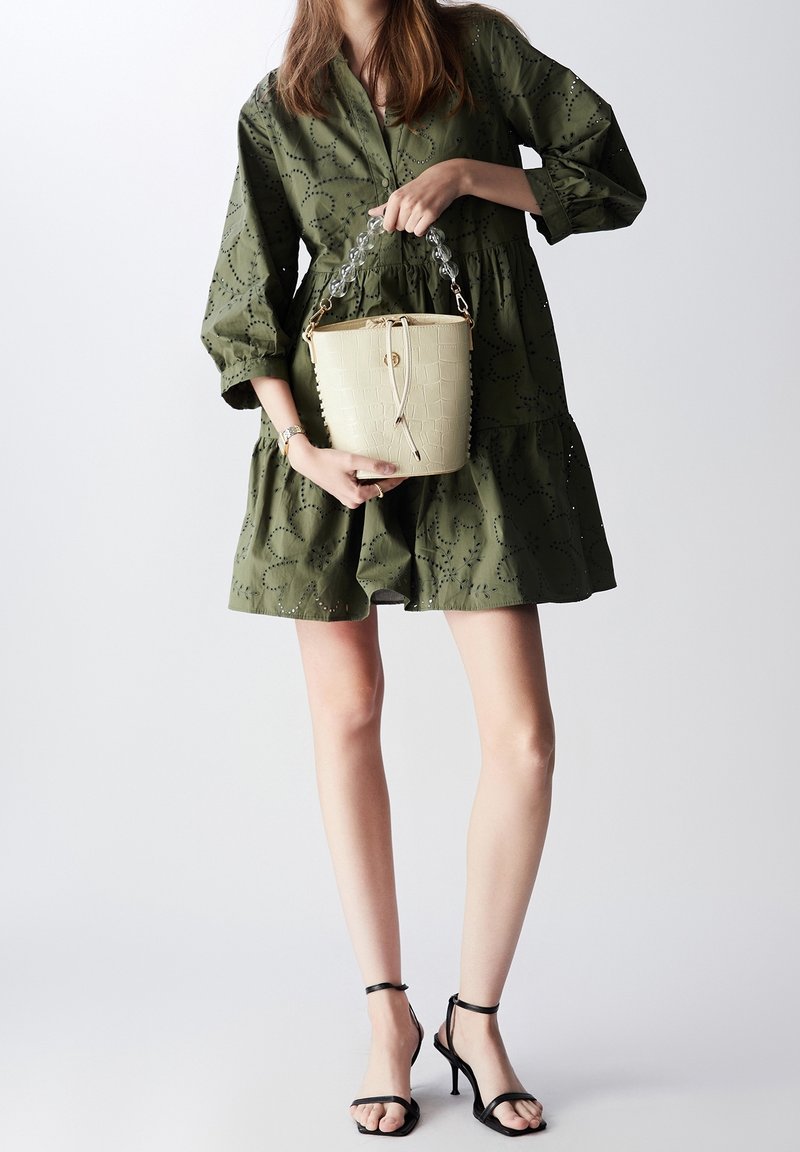 Femme en robe vert olive tenant un sac seau beige avec des anses en perles transparentes, portant des sandales noires à talons hauts.