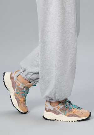 Piedi con sneakers multicolori con accenti beige, marrone e verde acqua abbinati a pantaloni della tuta grigio chiaro su uno sfondo grigio.