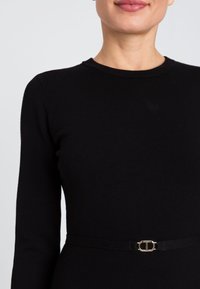 Abito maglione nero con scollatura rotonda, maniche lunghe e vita aderente. Presenta una cintura nera con dettagli in oro.