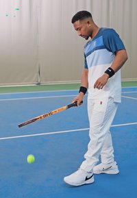 Mann i blå og hvit sportsklær som sprett en tennisball med et trek-trekk over en innendørs blå bane.