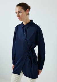 Cămașă albastră închisă, cu nasturi, guler, design înfășurat și legătură pe lateral. Mâneci lungi și textură fină. Se asortează cu pantaloni de culori deschise.