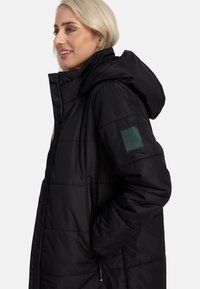 Melnā izolētā jaka ar kapuci, ar quilted dizainu, ar rāvējslēdzēja kabatām un zaļu logo uz kreisā piedurknes. Gluda tekstūra.