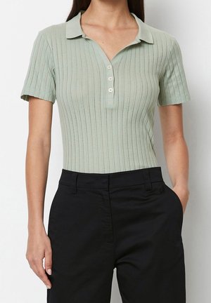 Kvinde iført en lys grøn ribbet kortærmet poloshirt stoppet ned i højtaljede sorte bukser med den ene hånd i lommen.