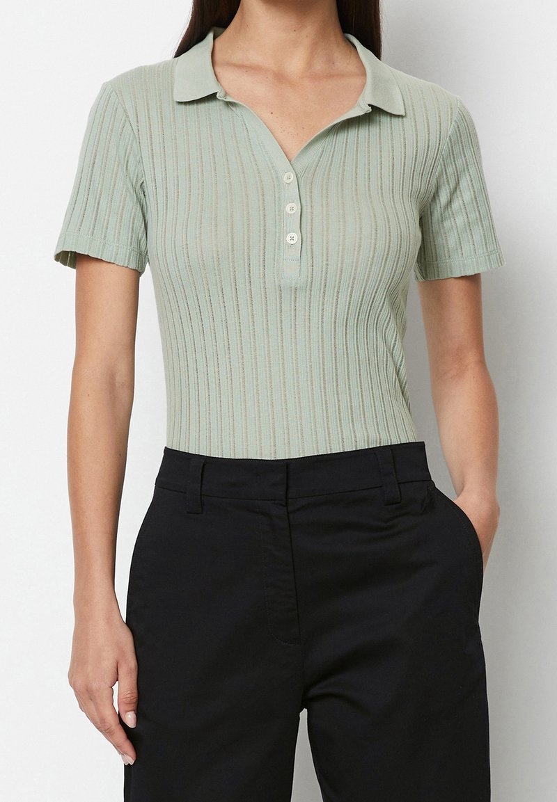 Femme portant un polo côtelé vert clair à manches courtes, rentré dans un pantalon noir taille haute, avec une main dans la poche.