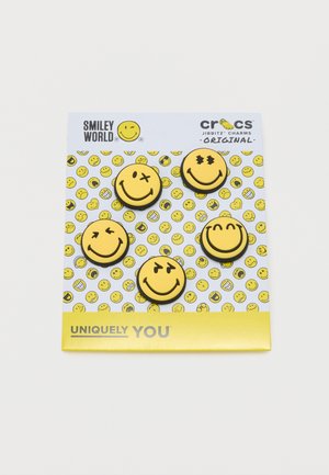 Five yellow smiley face Crocs Jibbitz charms with different expressions on a white card with scattered smiley faces background.