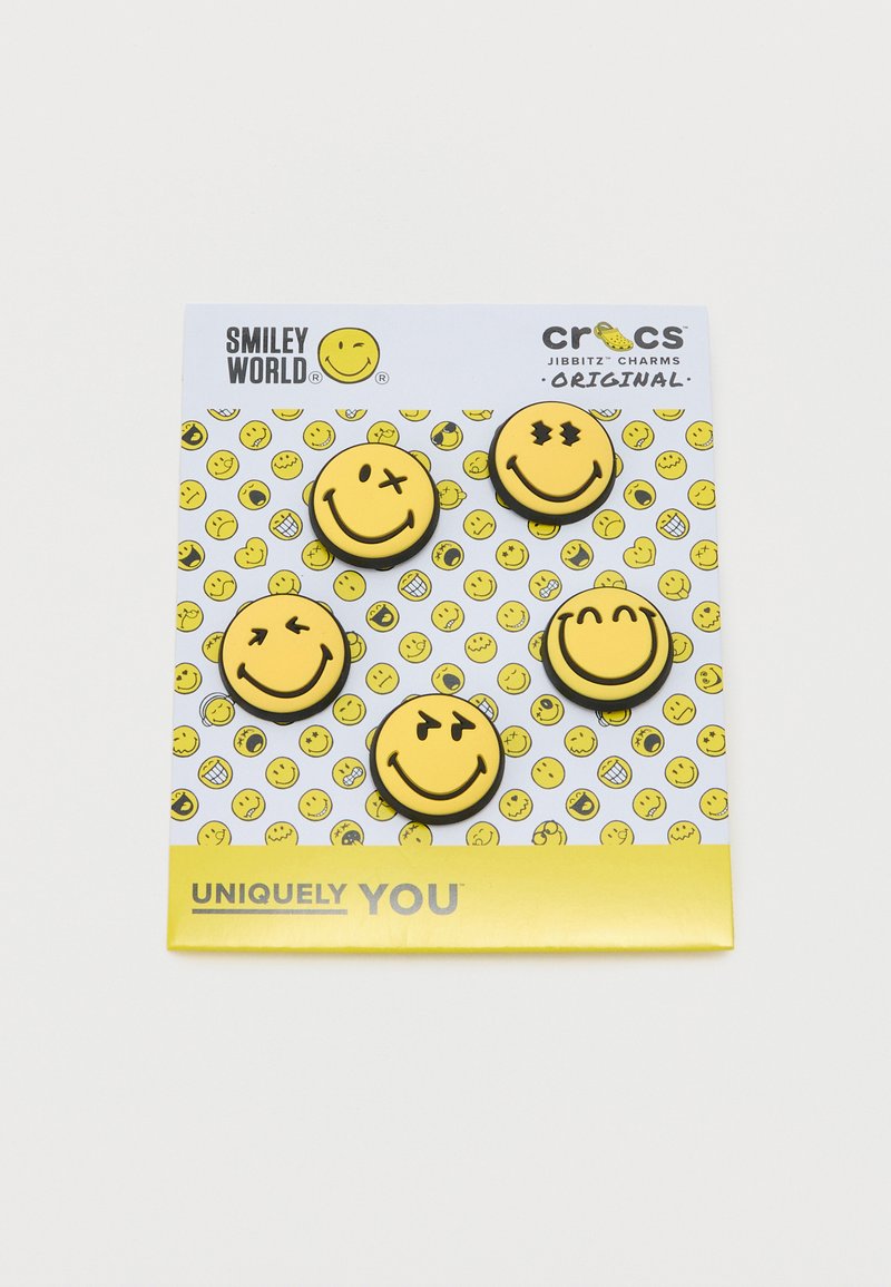 Πέντε κίτρινα γκρινιάρικα Jibbitz Crocs με διαφορετικές εκφράσεις σε μια λευκή κάρτα με φόντο από διασκορπισμένα γκρινιάρικα πρόσωπα.