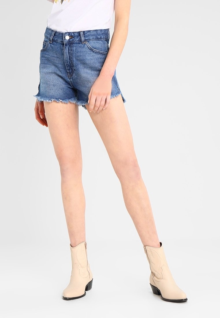 Farkkushortsit, joissa on repaleinen helma, korkea vyötärö ja vaaleansininen väri, yhdistettynä beigeklassisiin nilkkabootseihin, joissa on koristeellisia tikkejä.