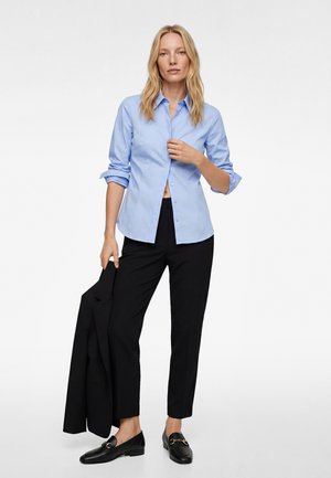 Frau steht und trägt ein hellblaues Hemd mit Knöpfen, schwarze verkürzte Hosen, schwarze Loafers und hält einen schwarzen Blazer in ihrer linken Hand.