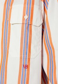 Randig skjorta med orange och blå vertikala ränder på en krämfärgad bakgrund, med bröstficka och broderade röda initialer.