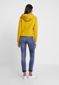 Sudadera amarilla con capucha ajustable, combinada con jeans skinny azules con dobladillo deshilachado. Usada con zapatillas blancas, vista desde atrás.
