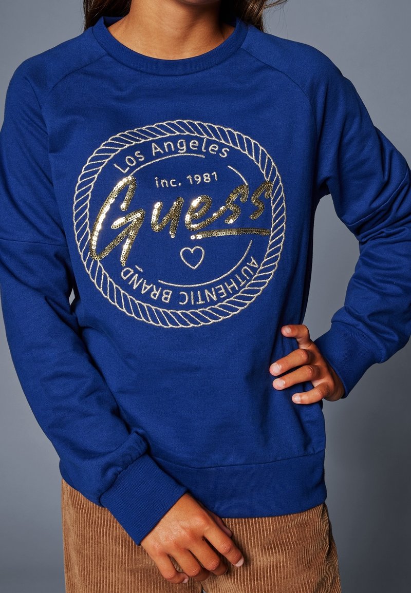 Blå sweatshirt i bomullsblandning med ribbade ärmar. Har en logotypdesign i guldglitter med repdetaljer och tryckt text.