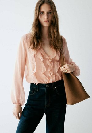 Joven mujer con una blusa rosa transparente con volantes, vaqueros oscuros, llevando un gran bolso marrón al hombro y con dos anillos en la mano derecha.