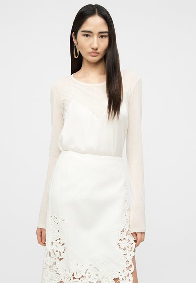 Γυναίκα με μακριά σκούρα μαλλιά φοράει ένα διαφανές τοπ με μακριά μανίκια κάτω από ένα λευκό φόρεμα χωρίς τιράντες και μια λευκή φούστα με λουλουδάτες δαντελένιες κοψίματα.