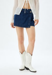 Jupe mini en denim avec un design enroulé, dotée d'une fermeture à bouton, de couleur bleu foncé et d'un ourlet surpiqué. Portée avec des bottes de cow-boy blanches.