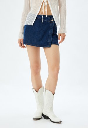 Jupe mini en denim avec un design enroulé, dotée d'une fermeture à bouton, de couleur bleu foncé et d'un ourlet surpiqué. Portée avec des bottes de cow-boy blanches.