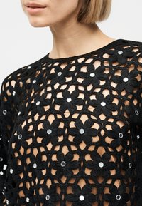 Femme portant un haut en crochet noir avec des motifs floraux ouverts et de petites décorations rondes argentées sur les fleurs.