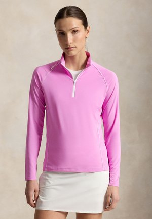 Giovane donna con capelli castani raccolti in uno chignon, che indossa una maglia rosa a maniche lunghe con zip e una gonna bianca su sfondo neutro.