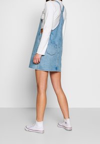 Denim overall dressi, vaaleansininen, hihaton ja vyötäröltä istuva. Ominaisuuksiin kuuluvat etutaskut, raaka helma, käytettynä valkoisten korkeiden tennarien kanssa.