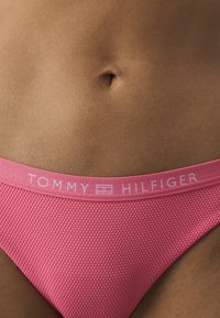 Rozā teksturēta bikini apakšdaļa ar platu logo jostasvītu, uz kuras ir uzraksts "TOMMY HILFIGER", ar gludu apdari un pieguļošu dizainu.
