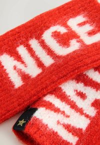 Bufanda de lana roja tejida con letras blancas gruesas que dicen "NICE", con una textura suave y esponjosa, y una pequeña etiqueta negra con un acento de estrella dorada.