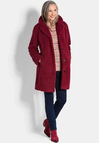 Frau, die lächelt und einen roten Wollmantel, einen mehrfarbigen Pullover, dunkelblaue Hosen und rote Stiefeletten trägt, vor einem schlichten Hintergrund steht.