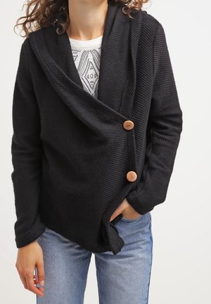Femme portant un cardigan noir texturé avec de gros boutons marron sur un t-shirt blanc imprimé et un jean bleu, main dans la poche.