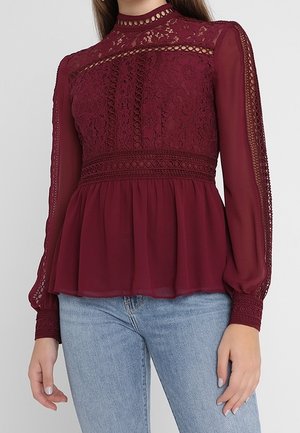 Mujer con blusa de encaje burdeos con cuello alto, mangas largas transparentes y bajo peplum combinada con vaqueros azul claro.