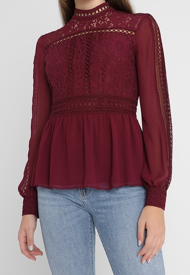 Femme portant un chemisier en dentelle bordeaux à col montant, manches longues transparentes et bas à basque, associé à un jean bleu clair.