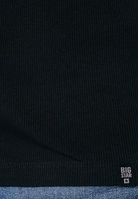 Juoda ribuota medžiaga su matomu siūlės kraštu virš mėlynos džinsinio audinio, kurioje yra mažas juodas etiketas su užrašu „BIG STAR“ ir žvaigždės piktograma.