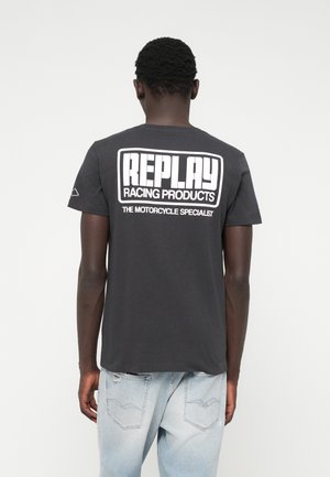 Czarna bawełniana koszulka z dużym białym logo "REPLAY RACING PRODUCTS" na plecach. Krótkie rękawy, okrągły dekolt, luźny krój, zestawiona z jasnoniebieskimi dżinsami.