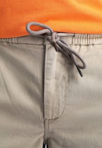 Beige broek met een elastische tailleband en een contrasterend grijze koord. Het koord heeft zwarte aglets en een gladde, matte textuur.