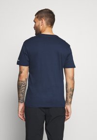 Navy blue cotton t-shirt with short sleeves, crew neckline, and seamless shoulder design. Features visible arm tattoos on the model.