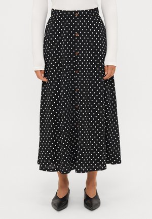 Zwarte midirok met witte polka dots en bruine knopen aan de voorkant, gedragen met een witte top met lange mouwen en zwarte schoenen met een puntige neus.