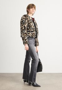 Jas van nepleer met luipaardprint en zwarte afwerking, gecombineerd met grijze flared jeans met rafelige zoom en zwart puntige enkellaarsjes, terwijl ze een zwarte tas vasthoudt.