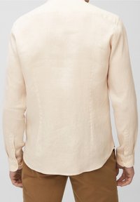 Uomo che indossa una camicia di lino beige a maniche lunghe e pantaloni marroni, mostrato da dietro su uno sfondo semplice.