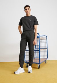 Sort bomulds sports skjorte og joggers med hvidt brandlogo, parret med hvide sneakers. Baggrunden har en blå metalvogn på guldtgulv.