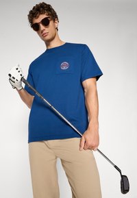Blå bomull t-skjorte med brystlomme og et rundt logo, sammen med beigefargede bukser. Modellen holder en golfkølle iført hvite hansker og solbriller.