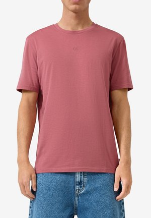 Man draagt een effen stoffig roze T-shirt met korte mouwen en ronde hals en blauwe spijkerbroek, staat met handen ontspannen langs het lichaam.