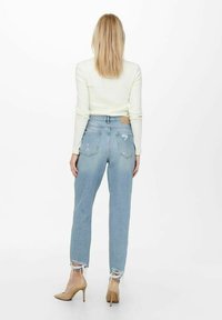 Vaaleansiniset korkeavyötäröiset farkut, joissa on rispaantuneet lahkeet, yhdistettynä istuvaan valkoiseen topiin ja beigeihin korkokenkiin. Takakuva, minimalistinen tausta.