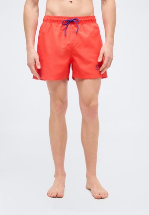 Mees, kes kannab erkpunaseid ujumispükse sinise nööriga ja vasakul reiel "EA" logoga, seisab paljajalu valge tausta ees.