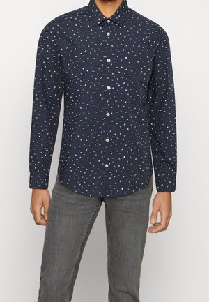 Marineblaues langärmliges Hemd mit kleinen weißen und braunen abstrakten Mustern, getragen zu grauen Jeans.