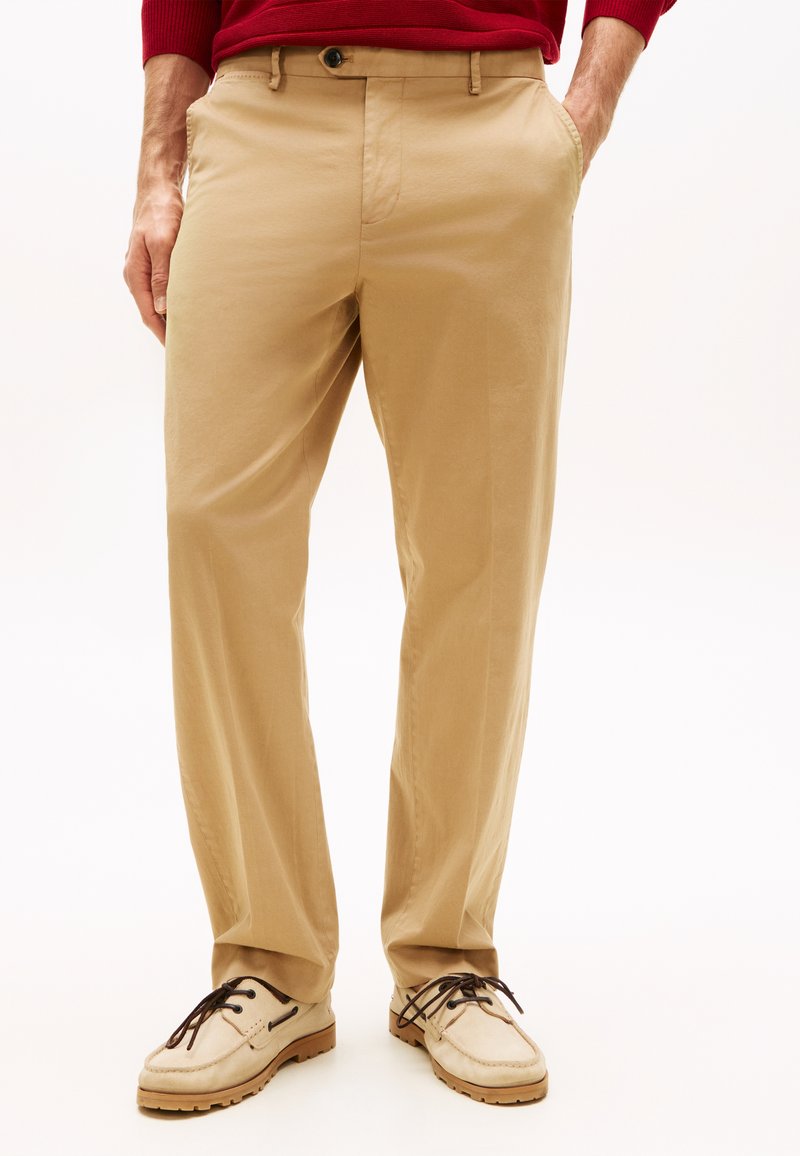 Άνδρας με μπεζ chinos, με τα χέρια στις τσέπες, συνδυασμένα με κρεμ casual παπούτσια και κόκκινο πουλόβερ ορατό στη μέση.