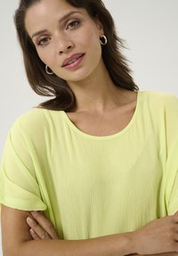 Top suelto de color amarillo claro con escote redondeado, tejido texturizado y mangas cortas. El modelo tiene los brazos cruzados y lleva pendientes de aro de plata.