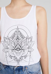 Débardeur en coton blanc avec un grand motif de fleur de lotus en contour noir, comportant des détails intriqués et des accents à pois. Coupe décontractée.