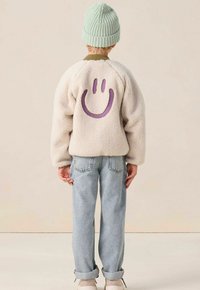 Kind met een beige fleece jack met een paars glimlach gezicht uitsnede, lichtblauwe jeans, beige sneakers en een lichtgroene gebreide muts, kijkend weg.