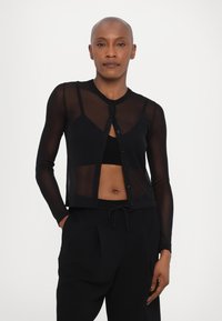 Black sheer cardigan with long sleeves, round neckline, and button front. Worn over a matching black top, it features a fitted design.