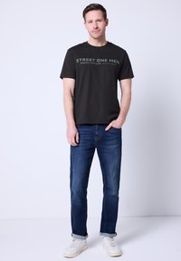 Czarny t-shirt z krótkim rękawem i zielonym napisem; zestawiony z niebieskimi jeansami i białymi tenisówkami. Prosty, casualowy krój.
